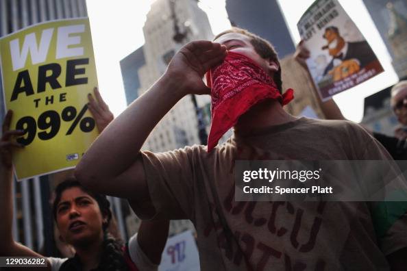 Getty images photo of a group of protestors with a young man in the extreme foreground in a red bandana. A woman to his right is holding up a placard reading WE ARE THE 99%