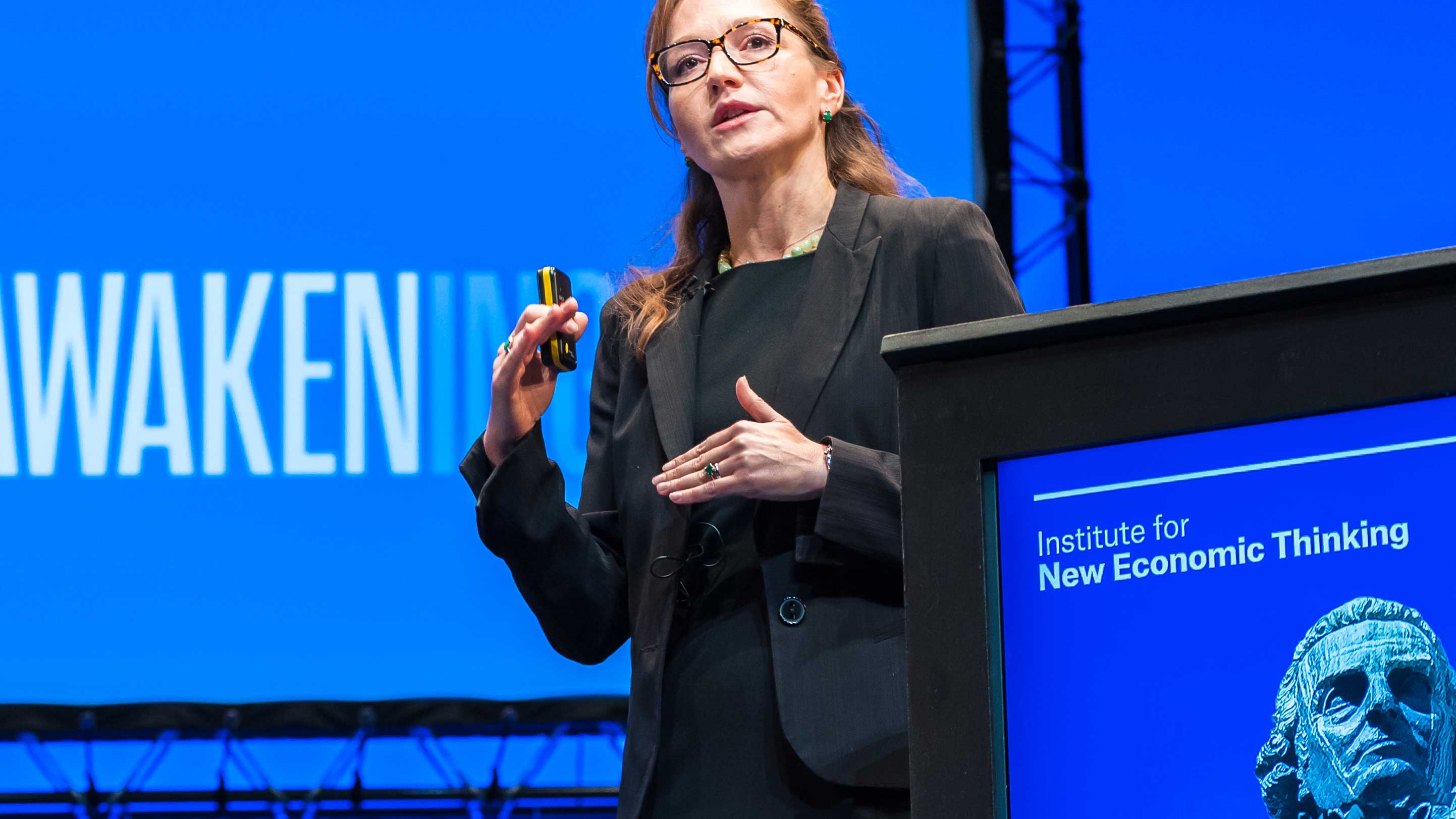 Anastasia Nesvetailova speaking at the Reawakening conference. Branded lectern is visible in the foreground and projected screen in the background.