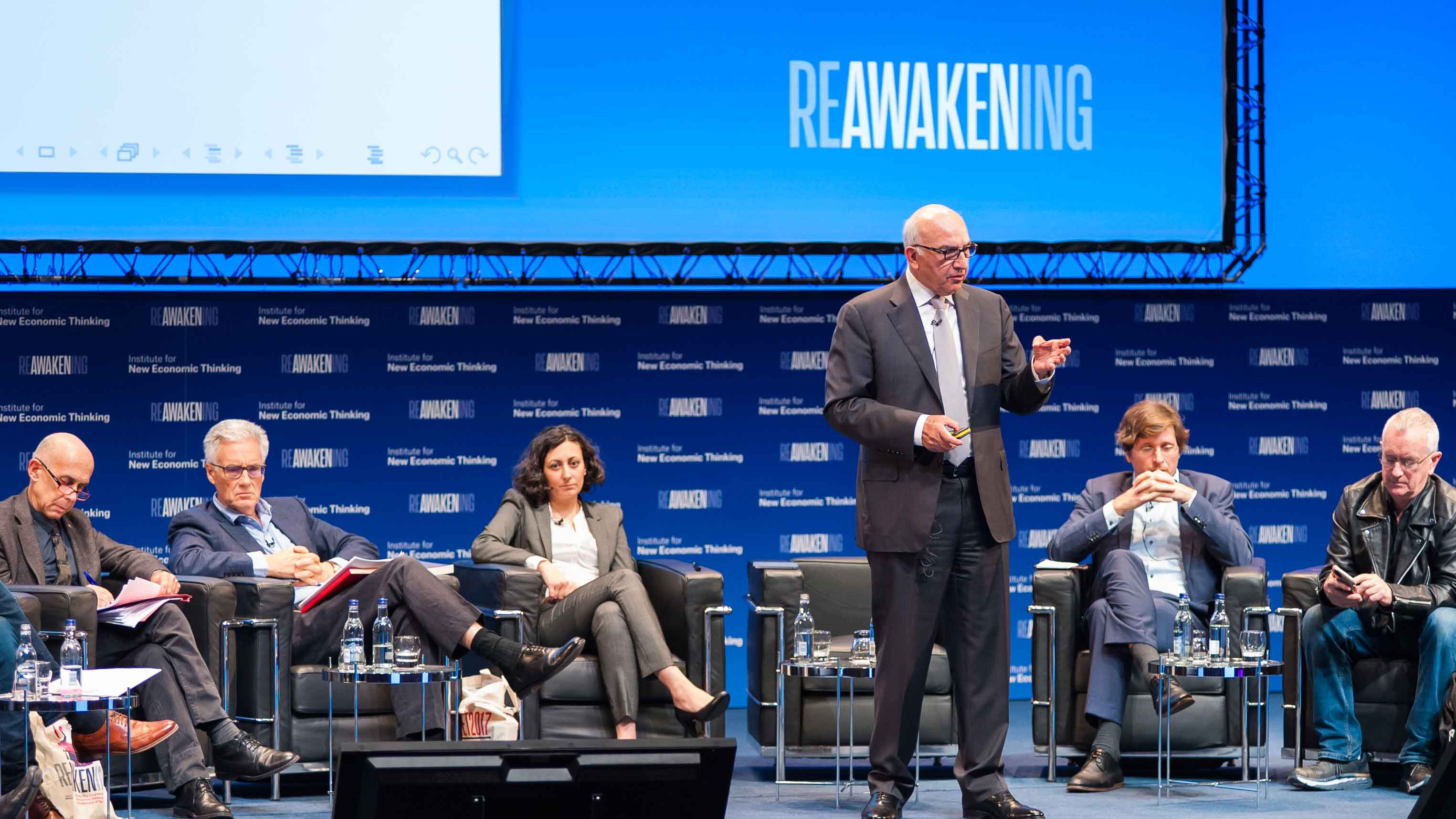 Hashem Pesaran speaking at the Reawakening conference as part of a panel group on stage. The background shows a branded confereence step-and-repear and projected backdrop.